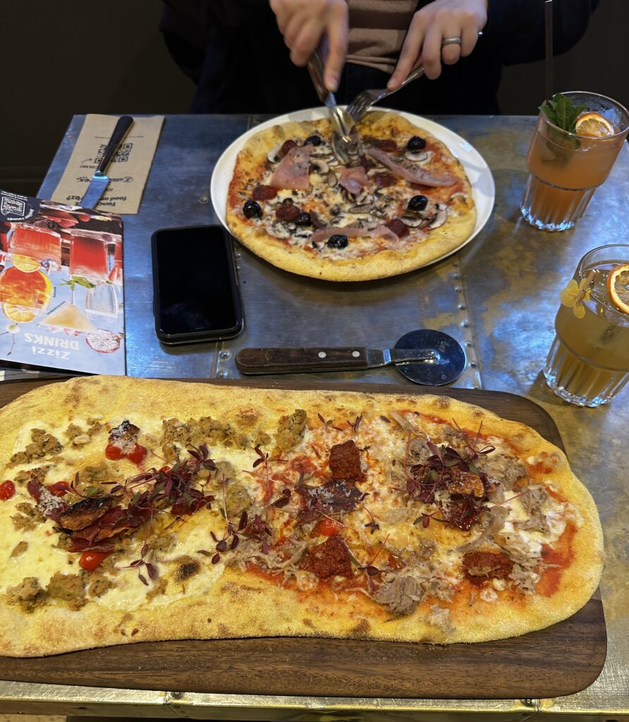 A table set with two pizzas, a pizza cutting wheel, and two drinks. The drinks are in glass with and orange slice floating on top and a yellow flower attached to the side. 